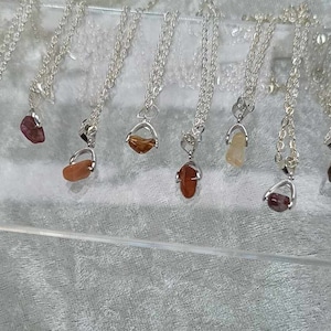 May include: A collection of eight silver necklaces with different colored gemstone pendants. The pendants are all in a silver setting and are shaped like a small, open cage. The gemstones are a variety of colors, including red, orange, yellow, brown, and blue.