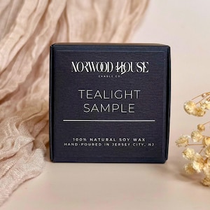 May include: A dark blue box with the text "NORWOOD HOUSE CANDLE CO" and "TEALIGHT SAMPLE" in white. The box also states "100% NATURAL SOY WAX HAND-POURED IN JERSEY CITY, NJ". The background includes a beige fabric and dried flowers.