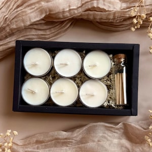 May include: A black box set containing six white tea light candles and a small glass bottle of matches. The candles are arranged in a grid, and the matches are next to them. The background includes a beige fabric and dried flowers.