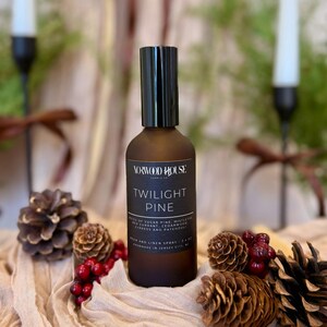 May include: A brown bottle of "Twilight Pine" room and linen spray with a black cap. The label lists ingredients like sugar pine and cedarwood. The bottle is surrounded by pine cones and red berries, with candles in the background.