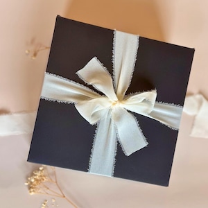 May include: A dark gray square gift box tied with a cream-colored ribbon and bow. The ribbon has a frayed edge, and the box is set against a soft pink background. The overall aesthetic is elegant and gift-ready.