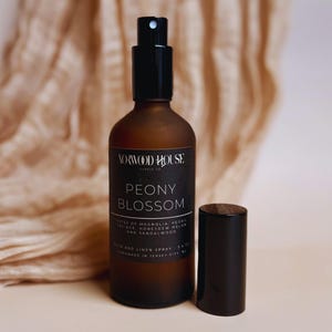 May include: A brown glass spray bottle with a black cap and a black label that reads "Peony Blossom." The bottle is next to a black cylindrical cap. The background is a beige fabric.