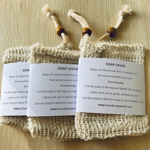 May include: Three beige soap saver bags made from natural sisal fibre, each with a drawstring closure and a wooden bead detail. Each bag has a white label with product information and the words "SOAP SAVER".