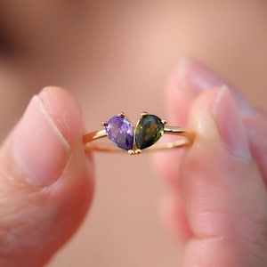 Puede incluir: Un anillo de oro con dos piedras preciosas en forma de lágrima. La piedra de la izquierda es morada y la piedra de la derecha es verde.