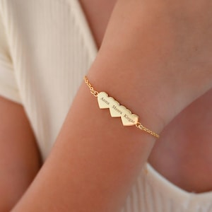 May include: A gold-colored bracelet featuring three heart-shaped charms. Each charm has an engraved name. The bracelet is worn on a wrist. The chain is delicate and the clasp is visible.