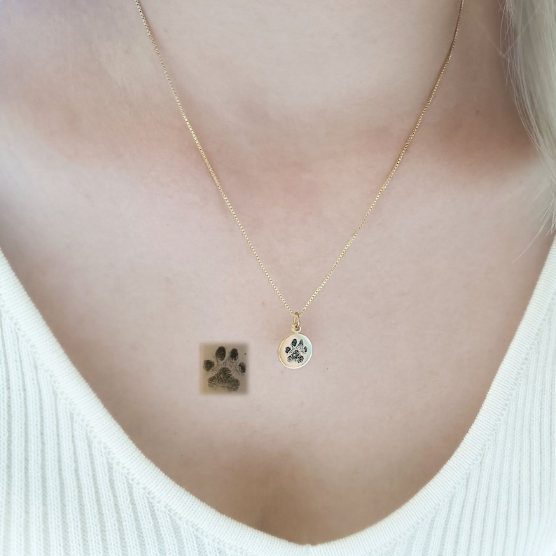 May include: A gold chain necklace with a round pendant featuring a black paw print. The pendant is worn by a person with a white shirt.