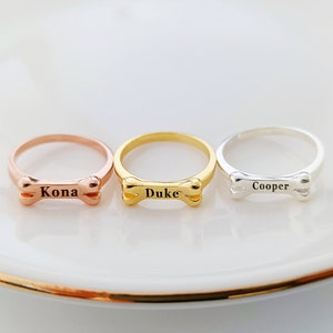 Puede incluir: Tres anillos de hueso de perro personalizados. Los anillos son de oro rosa, oro y plata. Los nombres "Kona", "Duke" y "Cooper" están grabados en los anillos.