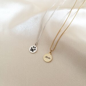 May include: Two silver necklaces with pendants. One pendant is a silver circle with a black paw print. The other pendant is a gold circle with the word "Meiko" engraved on it.