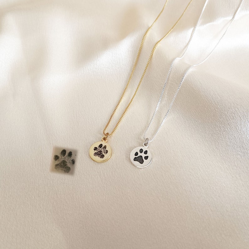 May include: Two necklaces with paw print pendants. One necklace has a gold pendant with a black paw print, the other has a silver pendant with a black paw print.