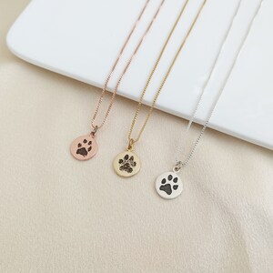 May include: Three silver necklaces with a paw print pendant. The necklaces are on a white surface. The paw prints are different sizes and are all black.