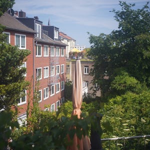 Puede incluir: Escena al aire libre con un edificio de ladrillo rojo con ventanas de marco blanco y un tejado oscuro. Una gran sombrilla beige cerrada se encuentra en primer plano, rodeada de exuberante follaje verde. El cielo es azul claro.