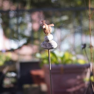Puede incluir: Una figurita de p&aacute;jaro de metal posada sobre una pila de tres piedras grises. El p&aacute;jaro es marr&oacute;n y tiene un pico largo y delgado. Las piedras est&aacute;n apiladas sobre una varilla de metal.