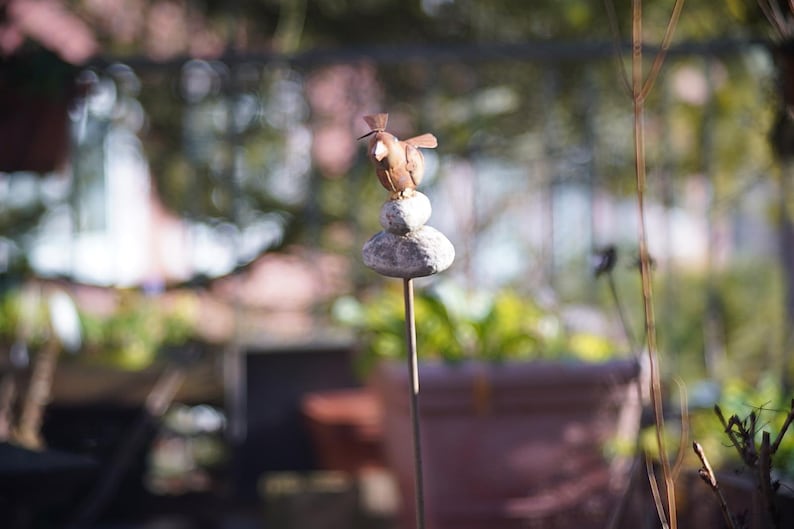 Puede incluir: Una figurita de p&aacute;jaro de metal posada sobre una pila de tres piedras grises. El p&aacute;jaro es marr&oacute;n y tiene un pico largo y delgado. Las piedras est&aacute;n apiladas sobre una varilla de metal.