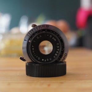 以下が含まれることがあります： 黒とシルバー仕上げのヴィンテージAgfa Karatカメラレンズ。レンズにはSolinar 1:3.5 5 cmのマーキングとRapidシャッタースピードダイヤルがあります。レンズは黒いベースに取り付けられ、木製の表面に置かれています。