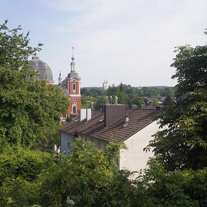 Puede incluir: Una vista exterior de una ciudad europea con una iglesia roja y blanca y un edificio con c&uacute;pula. Los edificios est&aacute;n rodeados de exuberantes &aacute;rboles verdes bajo un cielo azul.
