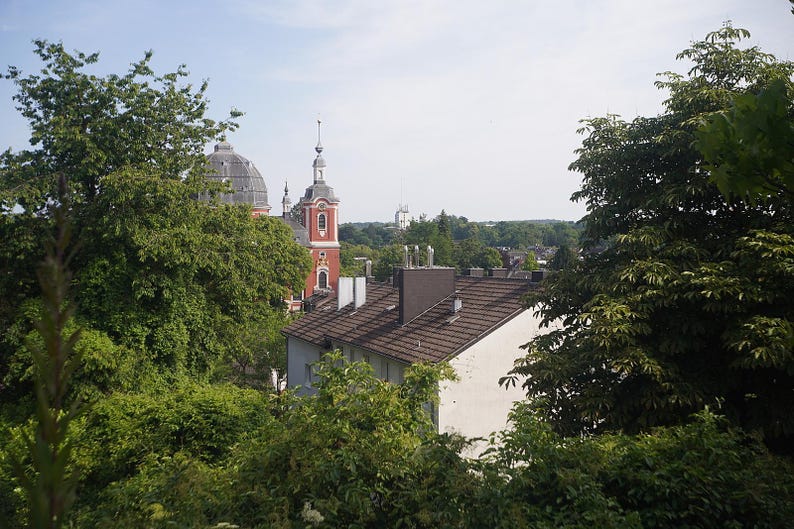 Puede incluir: Una vista exterior de una ciudad europea con una iglesia roja y blanca y un edificio con c&uacute;pula. Los edificios est&aacute;n rodeados de exuberantes &aacute;rboles verdes bajo un cielo azul.