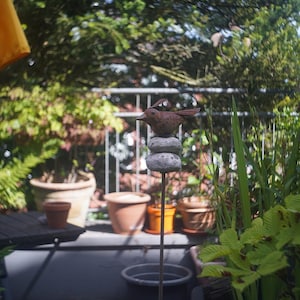 Puede incluir: Una escultura decorativa de un p&aacute;jaro de metal marr&oacute;n, posada sobre piedras apiladas, sobre un fondo de plantas en macetas y vegetaci&oacute;n. La escultura est&aacute; sobre una varilla de metal. La escena es al aire libre, con una sombrilla amarilla visible.
