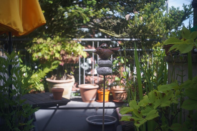 Puede incluir: Una escultura decorativa de un p&aacute;jaro de metal marr&oacute;n, posada sobre piedras apiladas, sobre un fondo de plantas en macetas y vegetaci&oacute;n. La escultura est&aacute; sobre una varilla de metal. La escena es al aire libre, con una sombrilla amarilla visible.