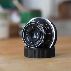 Puede incluir: Un objetivo de c&aacute;mara negro y plateado con marcas blancas, apoyado sobre una base negra. La lente tiene un elemento de vidrio transparente y se encuentra sobre un fondo borroso. Es un modelo T-43.