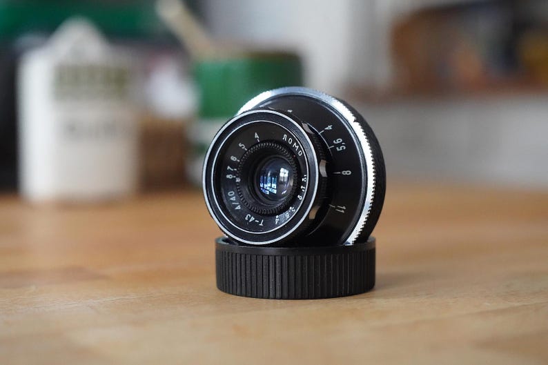 Puede incluir: Un objetivo de c&aacute;mara negro y plateado con marcas blancas, apoyado sobre una base negra. La lente tiene un elemento de vidrio transparente y se encuentra sobre un fondo borroso. Es un modelo T-43.