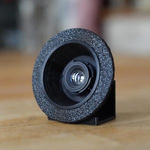 Op de afbeelding: Een zwarte ronde camera-lensadapter met een getextureerde, gespikkelde buitenrand en een gladde binnenbehuizing. Het apparaat is gemonteerd op een kleine, zwarte, L-vormige standaard, waardoor de lens verticaal wordt gepositioneerd.