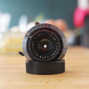 以下が含まれることがあります： ソリナー1:3.5レンズを備えたヴィンテージAgfa Karatカメラレンズ。レンズは黒とシルバーの仕上げで、黒いベースに取り付けられています。レンズの焦点距離は5cmで、シリアル番号は737686です。