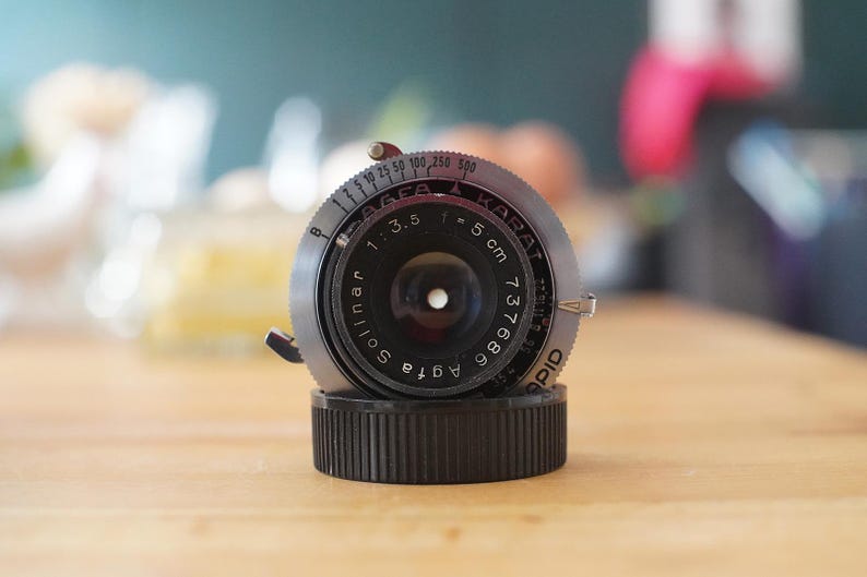 以下が含まれることがあります： ソリナー1:3.5レンズを備えたヴィンテージAgfa Karatカメラレンズ。レンズは黒とシルバーの仕上げで、黒いベースに取り付けられています。レンズの焦点距離は5cmで、シリアル番号は737686です。