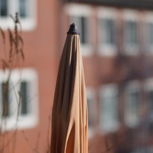 Puede incluir: Una sombrilla de patio beige con una punta negra, de pie frente a un fondo borroso de un edificio con ventanas.