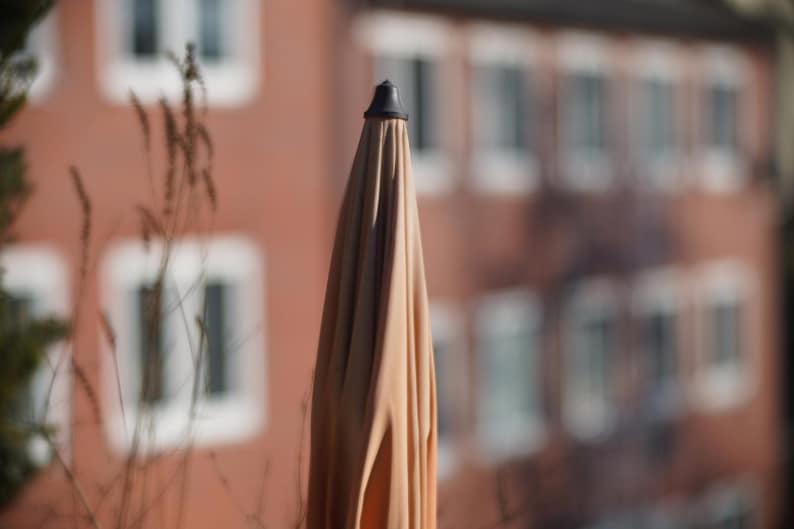 Puede incluir: Una sombrilla de patio beige con una punta negra, de pie frente a un fondo borroso de un edificio con ventanas.