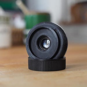 Puede incluir: Un objetivo de c&aacute;mara negro con un dise&ntilde;o circular, apoyado sobre una base a juego. El objetivo tiene una superficie reflectante y la base presenta una textura acanalada. El fondo est&aacute; borroso, con indicios de otros objetos.