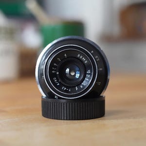 Puede incluir: Un objetivo de c&aacute;mara LOMO negro y plateado con una tapa de objetivo negra. El objetivo tiene marcas y n&uacute;meros alrededor del borde, incluyendo "LOMO" y "T-43". El objetivo est&aacute; sobre una superficie de madera.