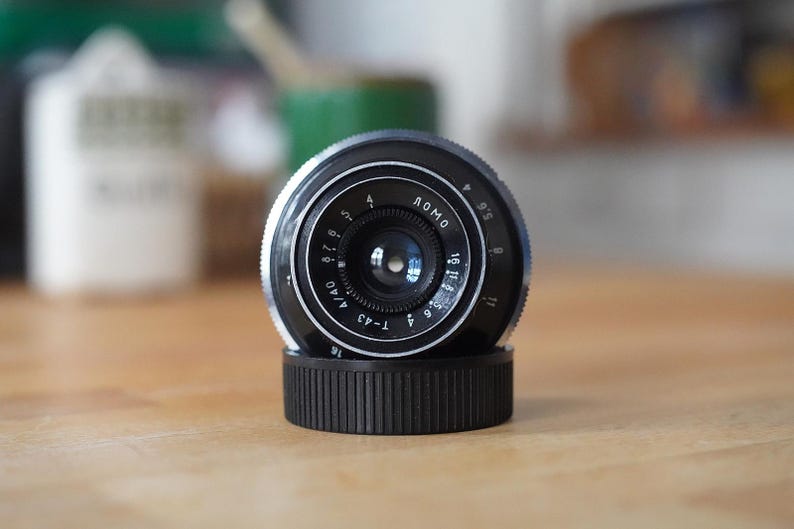 Puede incluir: Un objetivo de c&aacute;mara LOMO negro y plateado con una tapa de objetivo negra. El objetivo tiene marcas y n&uacute;meros alrededor del borde, incluyendo "LOMO" y "T-43". El objetivo est&aacute; sobre una superficie de madera.