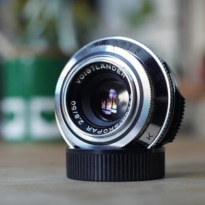 May include: A silver Voigtländer Color-Skopar 2.8/50 camera lens with a black ridged focus ring and aperture markings. The front element is clear, reflecting light, and the lens is resting on a black circular lens cap with a ribbed edge.