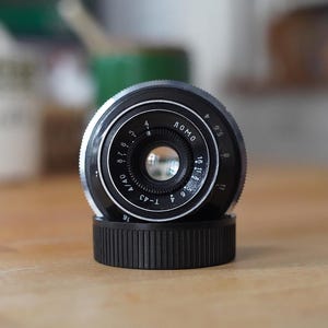 Puede incluir: Un objetivo de c&aacute;mara Lomo T-43 negro y plateado con una tapa de objetivo negra acanalada. La lente tiene marcas y n&uacute;meros blancos, incluyendo "LOMO" y "T-43 4/40". La lente est&aacute; sobre una superficie de madera.