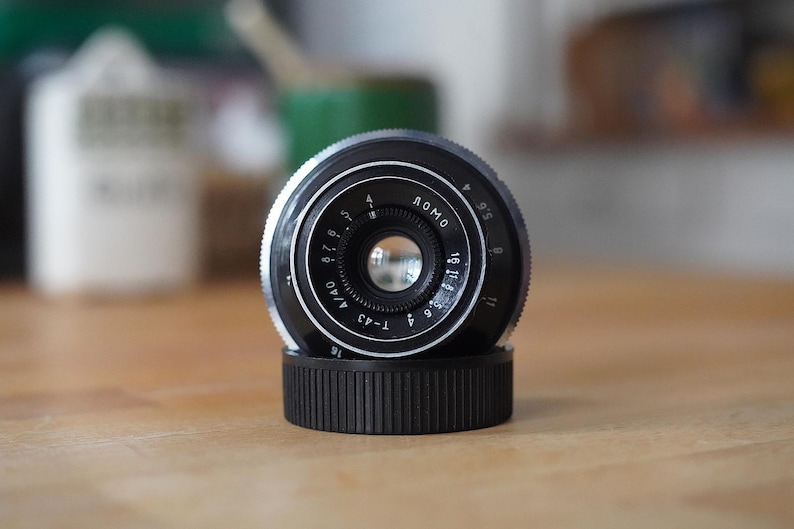 Puede incluir: Un objetivo de c&aacute;mara Lomo T-43 negro y plateado con una tapa de objetivo negra acanalada. La lente tiene marcas y n&uacute;meros blancos, incluyendo "LOMO" y "T-43 4/40". La lente est&aacute; sobre una superficie de madera.