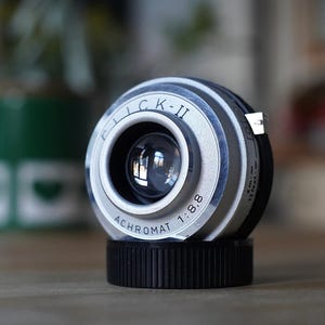 Op de afbeelding: Een zilverkleurige cameralens met een zwarte voet, voorzien van de tekst "FLICK-II" en "ACHROMAT 1:8.8" gegraveerd op de voorste ring. De lens heeft een centraal glaselement, een metalen lipje aan de zijkant en zichtbare afstandsmarkeringen.