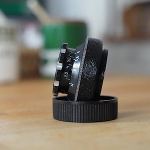 Puede incluir: Un objetivo de c&aacute;mara negro con marcas blancas y plateadas, apoyado sobre una tapa de objetivo negra. El objetivo tiene ajustes de apertura visibles, incluyendo f/2.8, f/4 y f/5.6. El objetivo est&aacute; colocado sobre una superficie de madera.