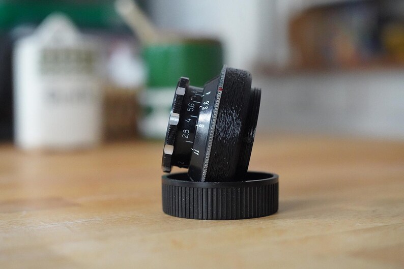 Puede incluir: Un objetivo de c&aacute;mara negro con marcas blancas y plateadas, apoyado sobre una tapa de objetivo negra. El objetivo tiene ajustes de apertura visibles, incluyendo f/2.8, f/4 y f/5.6. El objetivo est&aacute; colocado sobre una superficie de madera.