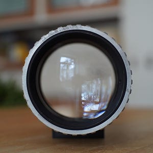 Puede incluir: Primer plano de un objetivo de c&aacute;mara plateado con un borde negro y una lente de vidrio transparente. El objetivo refleja una imagen borrosa de una ventana.