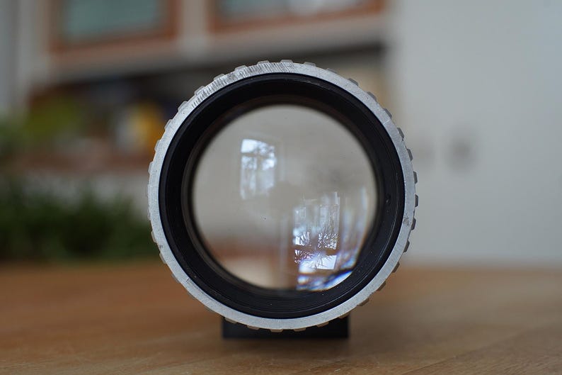 Puede incluir: Primer plano de un objetivo de c&aacute;mara plateado con un borde negro y una lente de vidrio transparente. El objetivo refleja una imagen borrosa de una ventana.