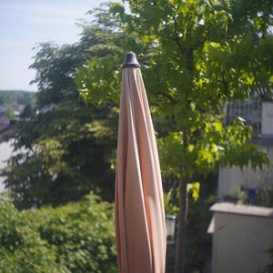 Puede incluir: Una sombrilla de patio cerrada de color melocot&oacute;n con una parte superior gris oscuro. La sombrilla est&aacute; rodeada de exuberantes &aacute;rboles y follaje verde, con un vistazo de cielo azul en el fondo. La sombrilla est&aacute; atada con tela a juego.