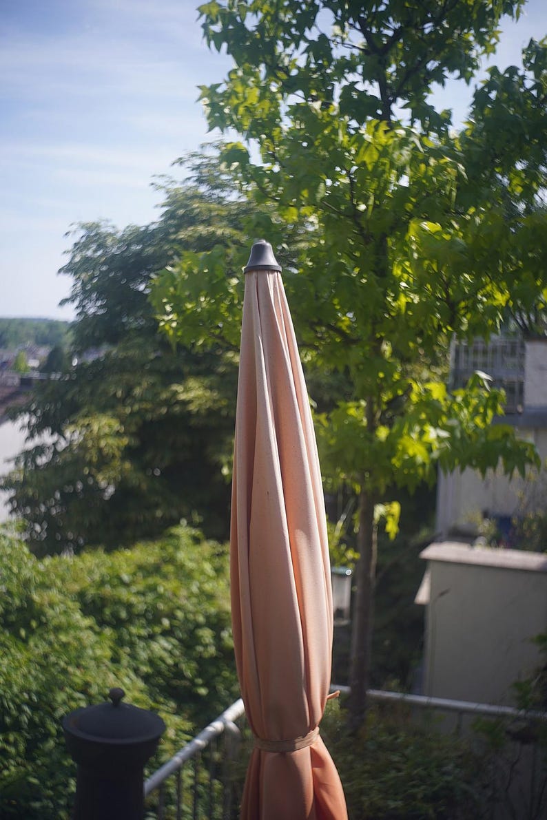 Puede incluir: Una sombrilla de patio cerrada de color melocot&oacute;n con una parte superior gris oscuro. La sombrilla est&aacute; rodeada de exuberantes &aacute;rboles y follaje verde, con un vistazo de cielo azul en el fondo. La sombrilla est&aacute; atada con tela a juego.