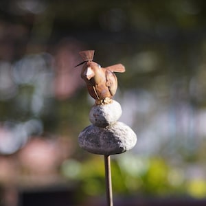 Puede incluir: Una escultura de p&aacute;jaro de metal oxidado posada sobre una pila de tres piedras grises. La escultura est&aacute; montada en una varilla de metal.