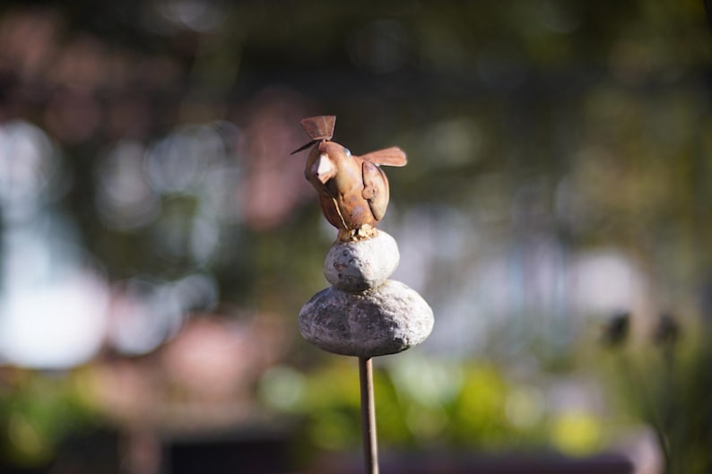 Puede incluir: Una escultura de p&aacute;jaro de metal oxidado posada sobre una pila de tres piedras grises. La escultura est&aacute; montada en una varilla de metal.