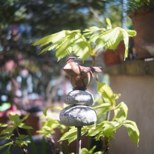 以下が含まれることがあります： 2つの積み重ねられた風化した石の上に止まっている茶色の鳥の彫刻が特徴の装飾的な庭の杭。鳥は翼を広げています。杭は緑の葉と鉢植えの植物に囲まれ、自然な屋外の美学を作り出しています。