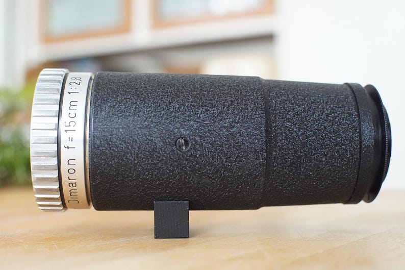 Puede incluir: Un objetivo de c&aacute;mara negro con una banda plateada que dice "Dimaron f=15cm 1:2.8".