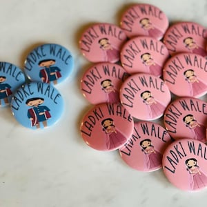May include: A collection of pink and blue round buttons with the text "Ladki Wale" and "Ladke Wale" printed on them. Each button features a cartoon illustration of a person in traditional Indian clothing.