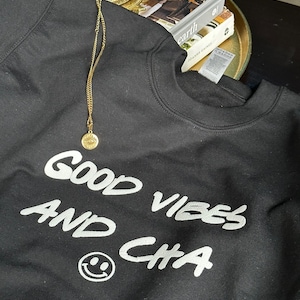 以下が含まれることがあります： 白い文字で「Good Vibes and Cha」と書いてある黒いスウェットシャツと白いスマイリー。スウェットシャツの上には、邪悪な目をモチーフにした金のネックレスが見える。