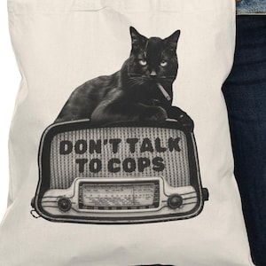 Puede incluir: Bolso tote color crema con un gato negro recostado sobre una radio vintage. El gato tiene un cigarrillo en la boca. La radio tiene las palabras "DON'T TALK TO COPS" impresas. El bolso tiene dos asas blancas.