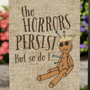 May include: A beige garden flag featuring the text "the HORRORS PERSIST But so do I..." and a cartoon voodoo doll. The flag is displayed on a black metal stand in a garden with green grass and vibrant flowers.